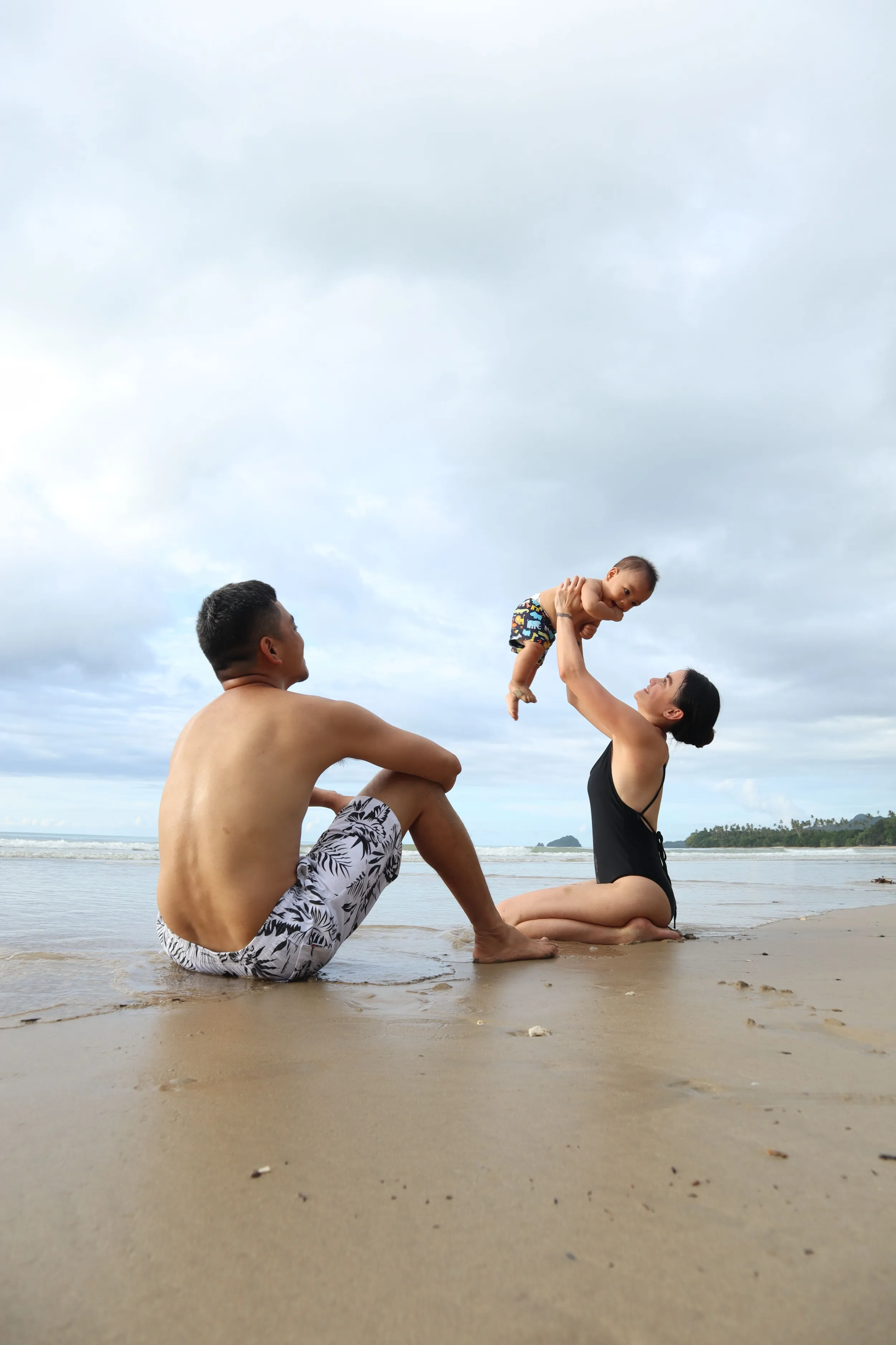 Mum lifting her baby into the air while dad relaxes on the sand at a Bali beach